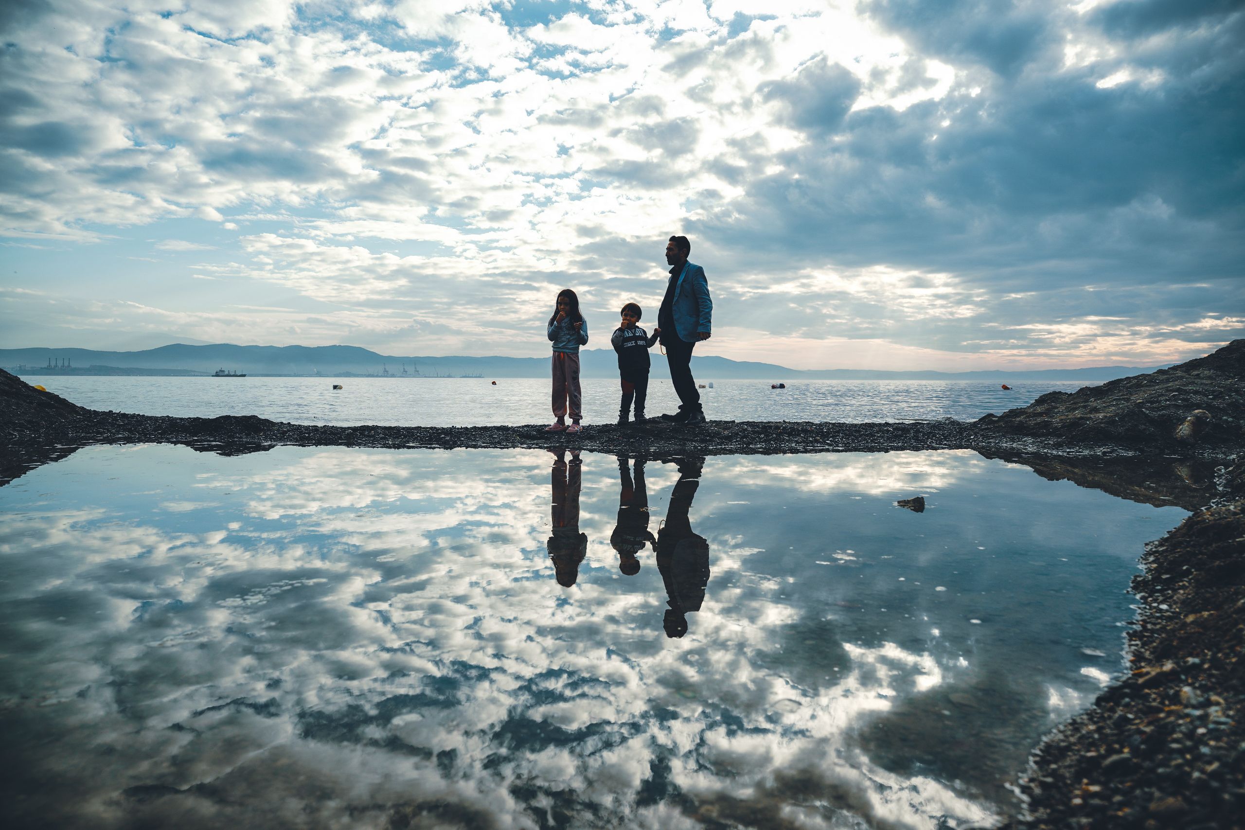 A serene family silhouette by the sea at twilight, reflecting in a tidal pool against a cloudy sky.